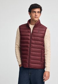 Chaleco de plumas burdeos con costuras verticales, cuello alto y cremallera frontal. Lleva puesto un suéter de punto crema y se combina con jeans oscuros.