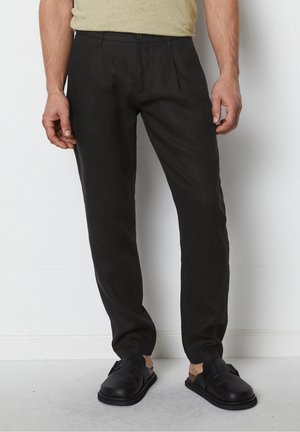 Mand iført slim fit sorte bukser, beige t-shirt og sorte slip-on sko, stående mod en ensfarvet hvid væg.