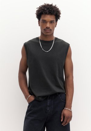 Junger Mann mit lockigem Haar, der ein ärmelloses schwarzes Hemd, eine silberne Kettenhalskette, Armbänder und dunkle Jeans trägt und mit einer Hand in der Tasche steht.