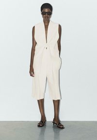 Crème-kleurige mouwloze jumpsuit met een strik in de taille, wijde pijpen en zijzakken; gecombineerd met zwarte zonnebril en zwart gespen sandaaltjes.