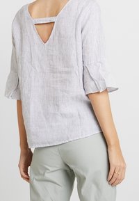 Žena ve světle šedé lněné blůze s rukávy do loktů a volánky, s V-zadním výstřihem a horizontálním popruhem, v kombinaci se světle šedými kalhotami.