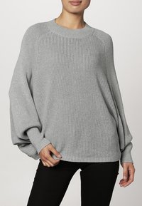 Světle šedý pletený svetr s širokou, žebrovanou strukturou, s velkorysými rukávy a kulatým výstřihem. V kombinaci s černými kalhotami.