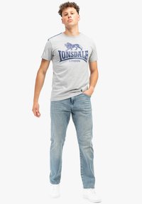 Pilkas trumpas rankovėmis marškinėliai su mėlynu liūto logotipu ir užrašu "Lonsdale London". Sujungta su šviesiai mėlynomis džinsais ir baltais sportbačiais.