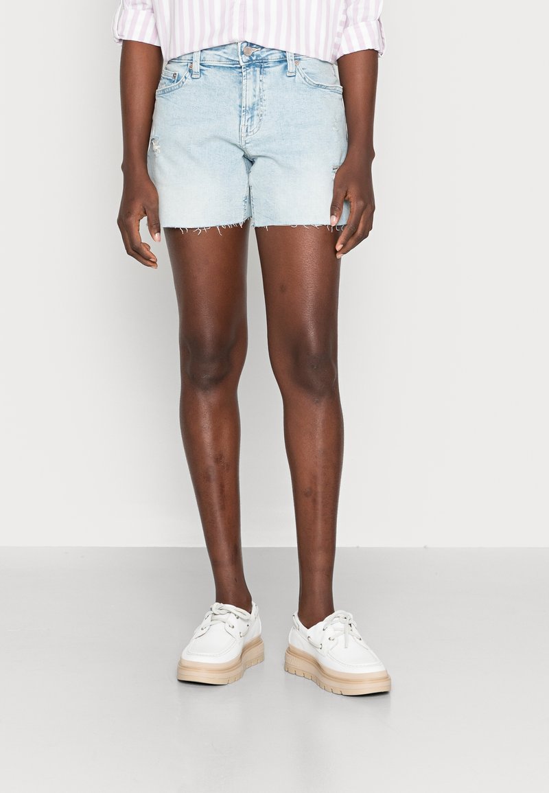 Ljusblå denimshorts med fransad nederkant, som har fem fickor. I kombination med vita snörskor på en enfärgad bakgrund.