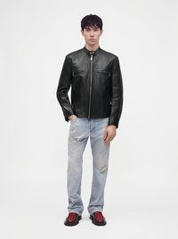 Jeune homme debout portant une veste en cuir noire, un jean clair déchiré et des chaussures noires avec des lacets rouges sur fond blanc.