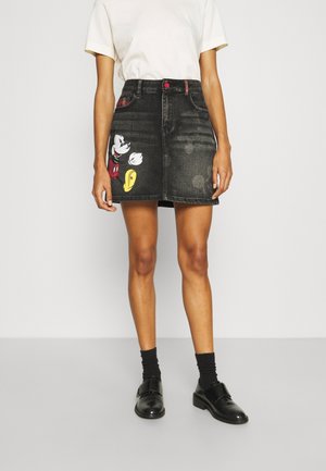 Vrouw die een zwarte spijkerrok draagt met een Mickey Mouse-print, een wit T-shirt, zwarte sokken en zwarte schoenen tegen een effen achtergrond.