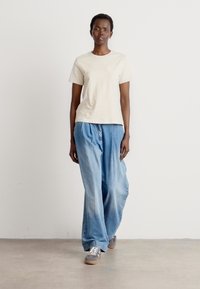 Beige puuvillane T-särk väikese taskuga, kombineeritud laia helesiniste denimipükstega. Hallid tennisetossud täiendavad riietust. Minimalistlik disain.