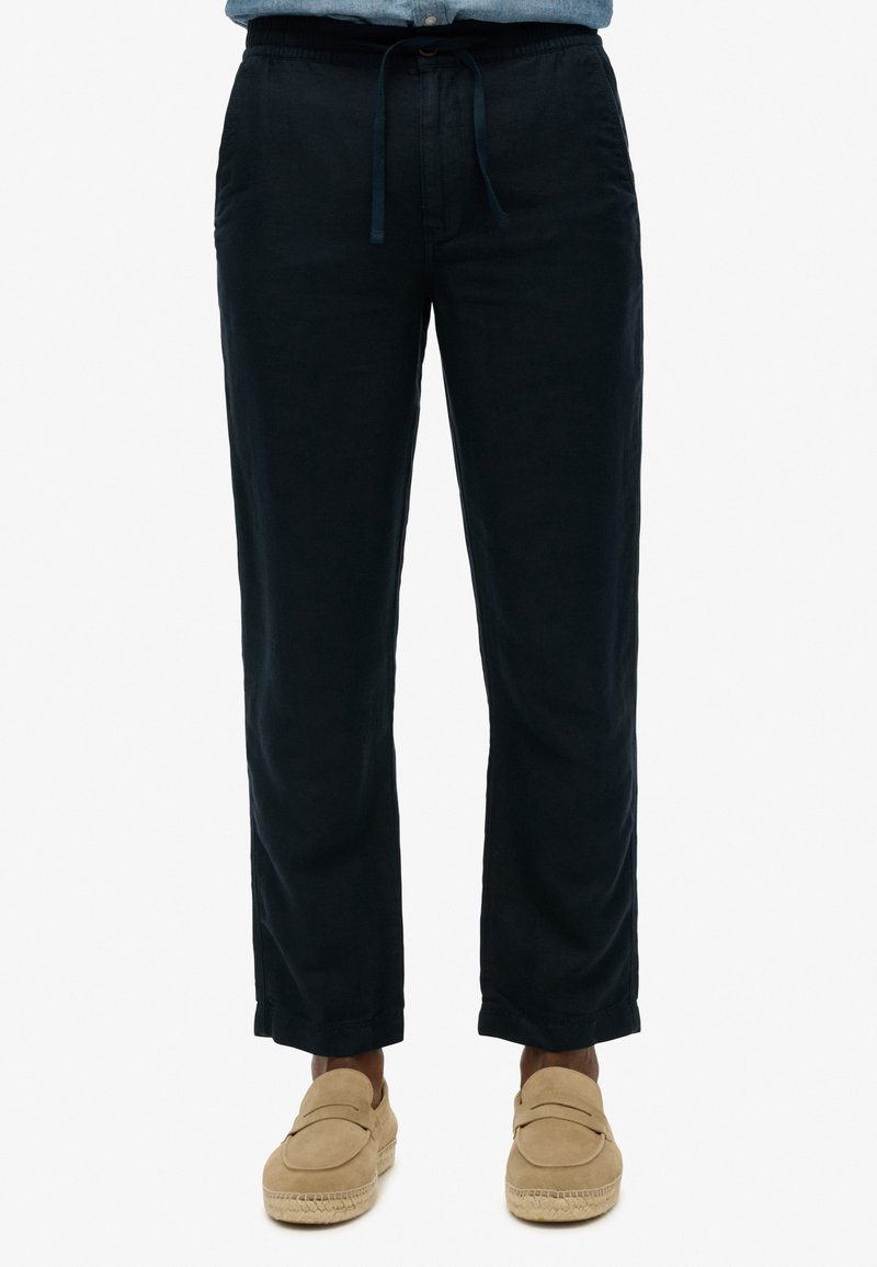 Homme portant un pantalon noir à cordon et des chaussures beige à enfiler, debout devant un fond uni.