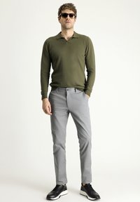 Polo a maniche lunghe verde oliva, pantaloni grigi slim-fit e scarpe nere con suole bianche. L'uomo sta dritto, indossando occhiali da sole.