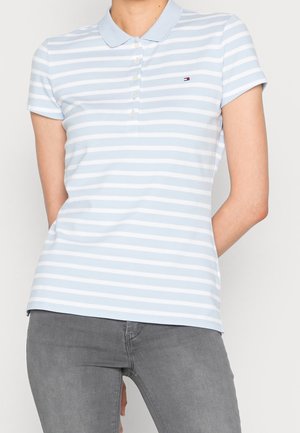 Henkilö, joka käyttää vaaleansinivalkoraidallista lyhythihaista poolopaitaa ja harmaita farkkuja, seisoo kädet selän takana.