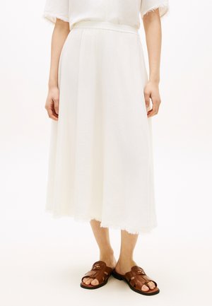 Femme portant une jupe midi blanche à ourlet brut et des sandales en cuir marron à bout ouvert, debout sur un fond blanc.