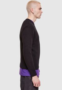 Pulover negru tricotat cu guler rotund, manșete și tiv cu nervuri, purtat peste un tricou violet. Expus din lateral pe un fundal neutru.
