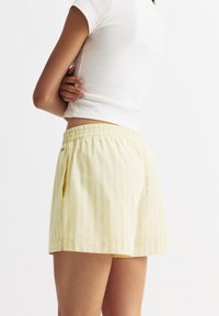 Hellgelbe Shorts mit vertikalen weißen Streifen, ausgestattet mit einem elastischen Bund, Seitentaschen und einem lockeren Schnitt. Hergestellt aus atmungsaktivem Material.