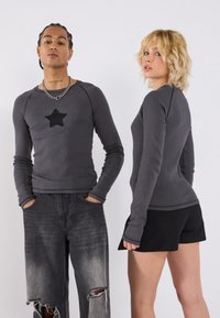 Hall grey pullover pika pikkade varrukatega, millel on must tähttikkimine rinnal, koos kulunud mustade teksade ja mustade lühikeste pükstega.