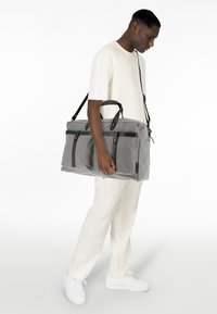 Sac de sport en tissu gris avec des accents en cuir noir, deux poches avant et deux poignées supérieures. Sangle d'épaule attachée.