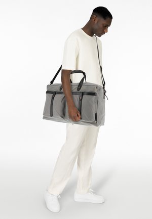 Bolso de lona gris con detalles en cuero negro, dos bolsillos frontales y dos asas superiores. Correa de hombro adjunta.