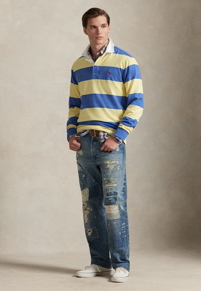 Πουκάμισο πόλο με ρίγες κίτρινου και μπλε χρώματος, μακρυμάνικο, με λευκό γιακά, σε συνδυασμό με ξεθωριασμένο τζιν με μπαλώματα και ελαφρώς φθαρμένο, ολοκληρωμένο με λευκά αθλητικά παπούτσια.