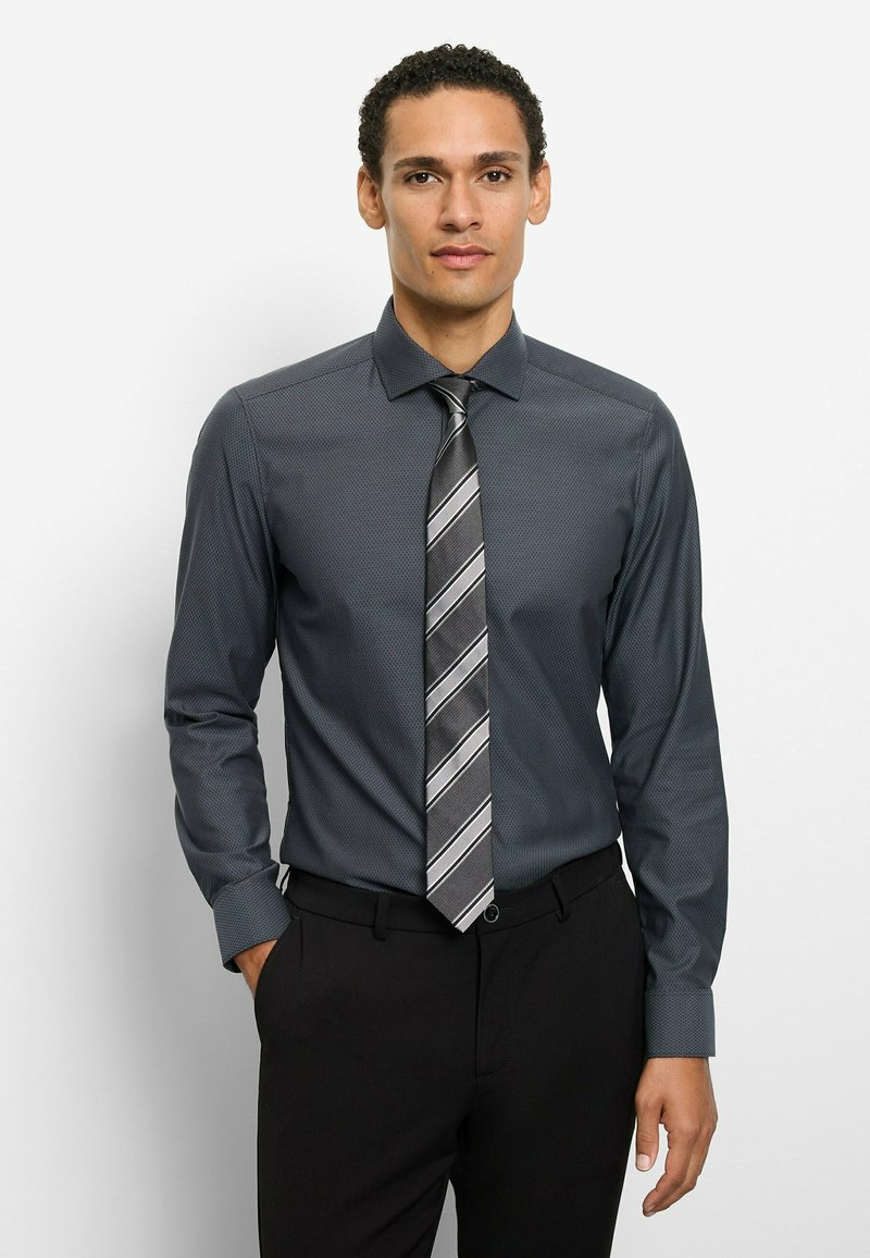 Dunkelgraues Hemd mit feinem Muster, Button-Down-Kragen und langen Ärmeln. Krawatte mit grauen und silbernen Streifen. Schwarze Hose.