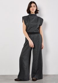 Grijze jumpsuit met een hoge kraag, een wijd lopende broek en een gestructureerde stof. Heeft een zwarte tailleband en korte mouwen, met een getailleerd silhouet.