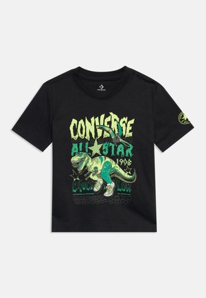 Zwart Converse T-shirt met korte mouwen, met een groene T-Rex die sneakers draagt en groene tekst met de woorden "Converse All Star Chuck Taylor 1908."