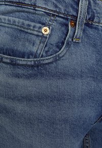 Vaqueros de mezclilla en azul medio con una textura sutil. Presentan costuras doradas y un botón metálico con el logotipo de la marca en el bolsillo delantero.