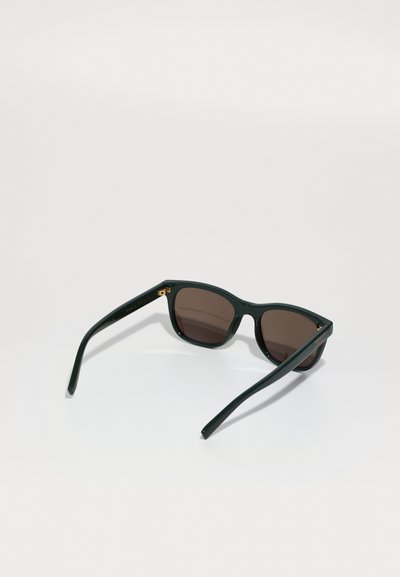 Groene plastic zonnebril met donkerbruine glazen, vierkant van vorm, en gouden accenten op het frame. Slanke armen steken uit het frame.
