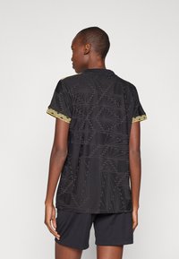 Svart sportsskjorte med geometrisk tekstur, korte ermer, og kontrasterende gul trim på mansjettene. Kombinert med svarte shorts.