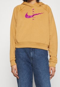 Hořčicově žlutá mikina s kapucí a růžovými logy Nike tištěnými vertikálně. Má volný střih a žebrované manžety a spodní lem. Viditelné denimbové džíny.