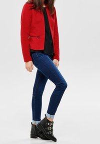 Rote, strukturierte Jacke mit Reißverschluss, kombiniert mit dunkelblauen Skinny-Jeans. Schwarze Knöchelstiefel mit drei Schnallenakzenten.