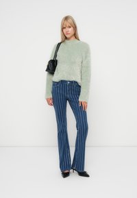 Hellgrüner, fusseliger Pullover mit langen Ärmeln, kombiniert mit blauen, ausgestellten Jeans mit weißen vertikalen Streifen. Kleine schwarze Tasche, die über die Schulter getragen wird.