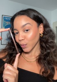 Brun ögonbrynspenna hålls mellan fingrarna, med en snygg design. Modellen bär en svart topp och lager av guldhalsband.