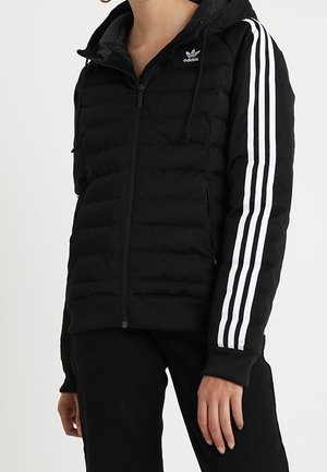Personne portant une doudoune noire Adidas avec trois bandes blanches sur les manches et une fermeture éclair à l'avant, associée à un pantalon noir.