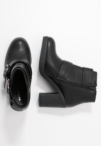 Schwarze Stiefeletten aus synthetischem Material, mit einem Reißverschluss, einem Riemen mit einem metallischen Akzent, runder Zehenpartie und einem klobigen, strukturierten Absatz.