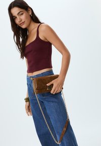 Pochette en daim marron avec une chaîne dorée, tenue d'une main. Le mannequin porte un crop top bordeaux foncé et un pantalon large bleu.