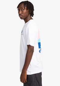 Camiseta de algodón blanca con diseño gráfico en azul y rosa en la parte posterior, que presenta el logotipo de la marca. Mangas cortas y corte relajado.