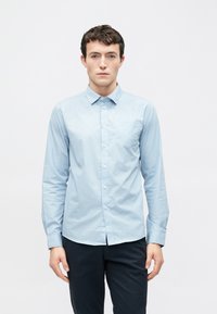 Chemise bleu clair à boutons avec un motif texturé subtil, col à revers, poche poitrine unique et manches longues. Portée avec un pantalon foncé.