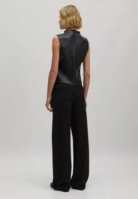 Zwarte mouwloze leren top met hoge kraag en rits aan de achterkant, gecombineerd met losse zwarte broek. Glad textuur en aangesloten ontwerp.