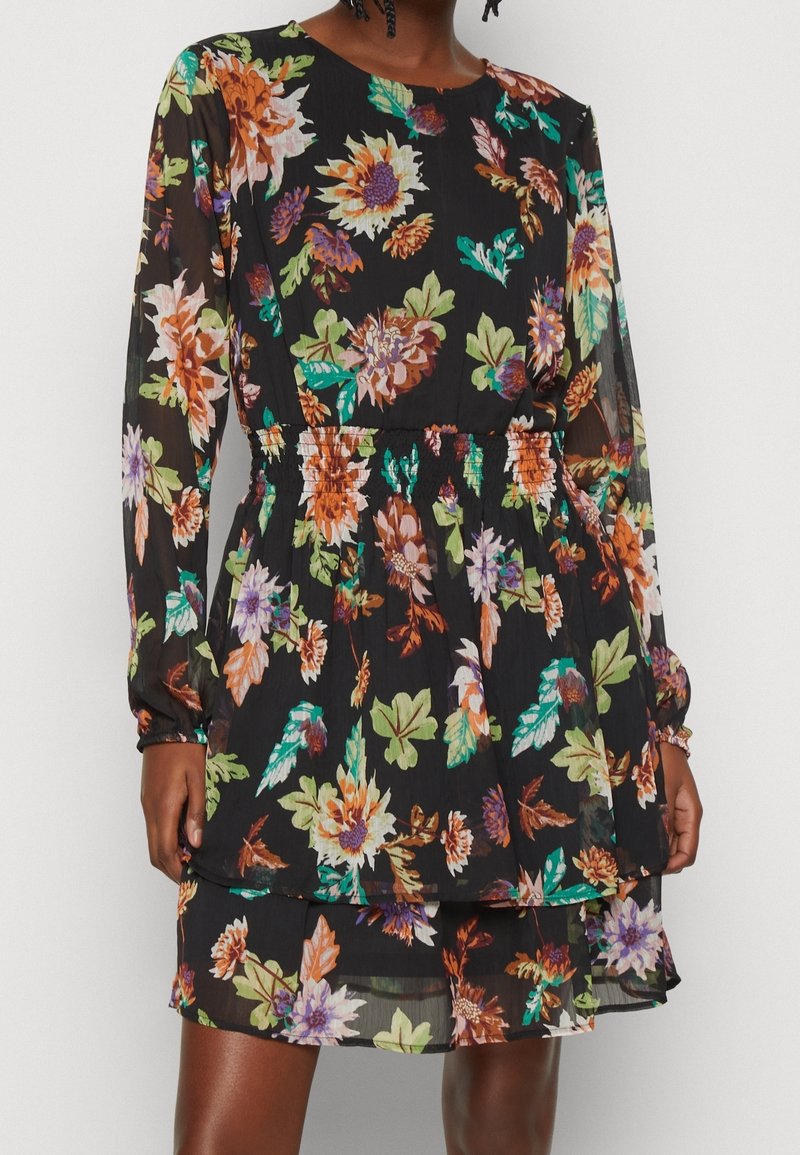 Robe longue à manches longues noire à fleurs orange, violettes et vertes, taille cintrée et jupe à volants portée par une personne.