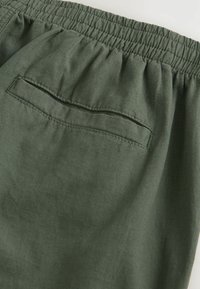 Elastisk talje shorts i mørkegrønt stof, med en enkelt baglomme med klap. Materialet er letvægts og tekstureret.