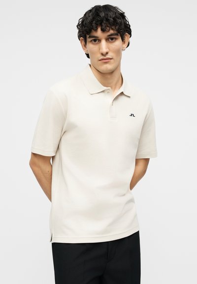 Νέος άντρας με σκούρα σγουρά μαλλιά, φορώντας μπεζ κοντομάνικο πόλο μπλουζάκι και μαύρο παντελόνι, στέκεται με τα χέρια πίσω από την πλάτη του.