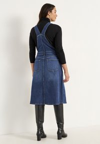 Denim hängselklänning i mörkblått med medellång längd. Utrustad med justerbara axelremmar, två bakfickor och en rak fåll.