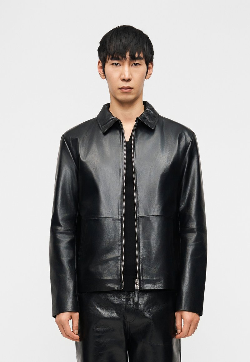 Νεαρός με ίσια μαύρα μαλλιά, φορώντας μια μαύρη δερμάτινη ζακέτα με φερμουάρ και ταιριαστό δερμάτινο παντελόνι, στέκεται μπροστά σε απλό φόντο.