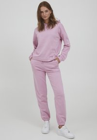 Helle rosa Kapuzenpullover mit lockerer Passform, kombiniert mit passenden Jogginghosen mit elastischem Bund und Bündchen. Weiße Sneakers runden das Outfit ab.
