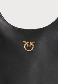 Saco de couro preto com um detalhe em metal dourado apresentando duas aves de frente uma para a outra em um anel circular.