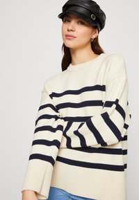Cream sweater with navy horizontal stripes, ribbed cuffs, and a round neckline. Features a relaxed fit and soft knit texture.