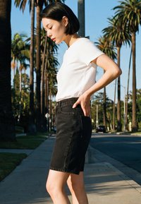 Fiatal nő fehér pólóban és fekete farmer szoknyában áll a járdán, mögötte pálmafák és autók, lefelé néz, kezét a csípőjén tartva.
