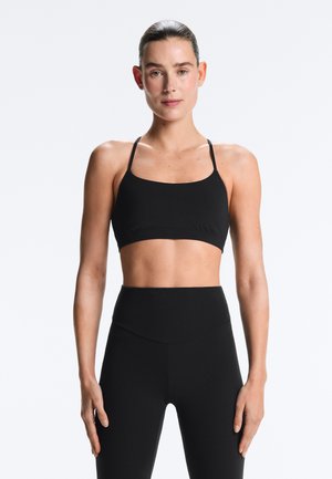 Vrouw met haar naar achteren gebonden, gekleed in een zwart sportbeha en high-waisted zwarte legging, staande tegen een effen lichte achtergrond.