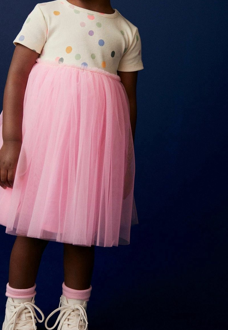 Enfant portant une chemise blanche à pois colorés, une jupe en tulle rose, des chaussettes roses et des baskets blanches sur un fond bleu foncé.