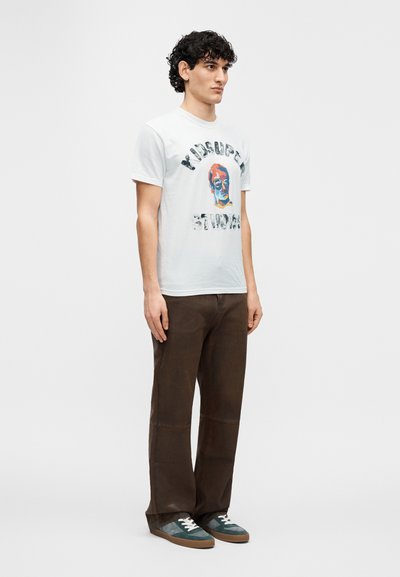 Λευκό κοντομάνικο μπλουζάκι με πολύχρωμο γραφικό τύπωμα και κείμενο, συνδυασμένο με καφέ παντελό νι με φαρδιά μπατζάκια και πράσινα αθλητικά παπούτσια με κορδόνια.