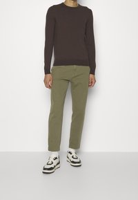 Pull en maille marron, pantalon vert olive et baskets noir et blanc. La tenue présente un col rond et une coupe fuselée dans un style minimaliste.